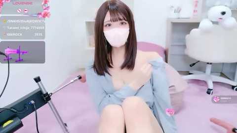 Media: A photo of an East Asian woman with shoulder-length brown hair, wearing a blue robe, pink face mask, and pink gloves, sitting on a pink cushion in a white room with a black chair.