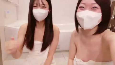 Video of two East Asian women with long black hair, wearing white face masks, towels around their waists, sitting on a tiled floor in a white-tiled bathroom.