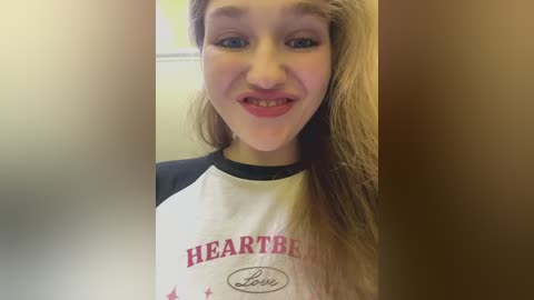 Video of a young woman with long blonde hair, light skin, and blue eyes, wearing a white and black heart-shaped shirt. She has a slight smile and a white substance on her face. Background is blurred.