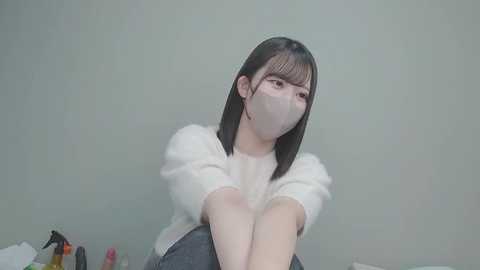 A video of an East Asian woman with straight black hair, wearing a white mask and a fluffy white sweater, sitting on a bed with a grey sheet. The background is a plain, light grey wall.
