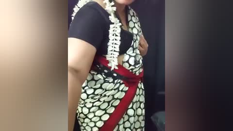 Video of a woman with medium skin tone, wearing a black short-sleeve top and a patterned sari with white polka dots, red borders, and intricate white lace detailing. The background is dark and blurred, with a slight focus on her body.
