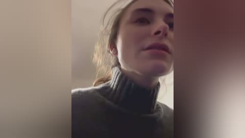A video of a young woman with fair skin, light brown hair, and light eyes, wearing a high-necked, dark gray sweater, gazing introspectively against a blurred, neutral background.