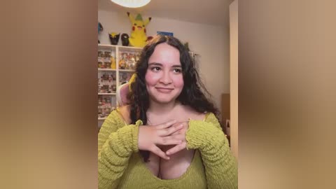Media: Video of a smiling woman with long, curly dark hair, wearing a mustard-yellow off-shoulder sweater, revealing her cleavage. Background features a Pikachu plush toy and a bookshelf filled with various items.