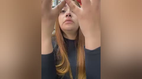 Video of a young woman with fair skin and long, straight auburn hair, wearing a black top. She is holding her hands up to her face, obscuring her features. The background is blurred, featuring a mix of colors, suggesting an indoor setting.