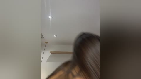 Media: Video of a blurred, nude woman with long, dark hair, viewed from behind. She stands in a minimalist room with white walls, recessed lights, and a wooden shelf. The image is slightly out of focus.