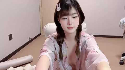 Video of a young East Asian woman with long black hair, wearing a pastel floral robe, sitting on a white cushioned chair in a minimalist room with beige walls and dark wooden floor.