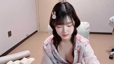 Video of a young Asian woman with fair skin and dark hair, wearing a pink floral robe, sitting on a beige chair in a minimalistic room with cream walls and a closed door.