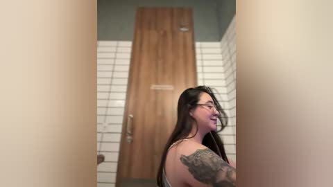 Video of a smiling woman with long black hair, glasses, and tattoos, standing in a narrow, tiled bathroom with a wooden door and green walls.