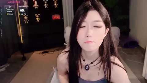 A video of an Asian woman with long black hair and fair skin, wearing a blue tank top and a necklace, sitting on a sofa in a dimly lit living room.