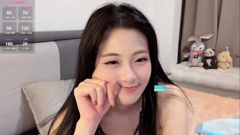 Video of an Asian woman with long black hair, wearing a black tank top, smiling with her hand on her cheek, in a bedroom with a gray headboard, stuffed toys, and a digital display.