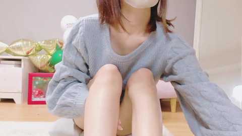 Media: A video of an Asian woman with straight, shoulder-length brown hair, wearing a loose, gray knitted sweater, sitting on a wooden floor in a cozy, softly lit room with a bed, green pillow, and a framed picture in the background.