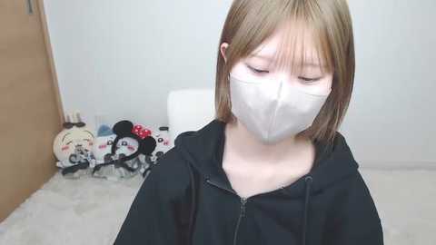 Video of a young Asian woman with light brown hair and pale skin, wearing a black hoodie and a white mask, standing in a minimalist room with a white wall, white carpet, and a wooden door.