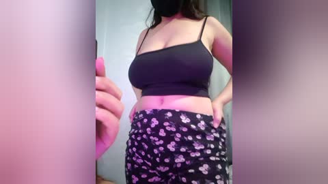 A video of a woman with light skin, wearing a black crop top and black pajama pants with a floral pattern. She has a medium build, small breasts, and a black face mask.