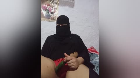 A video shows a middle-aged woman in a black niqab, red underwear, and holding a green dildo, seated on a bed with a patterned blanket. Background includes a wall and a basket of pink items.