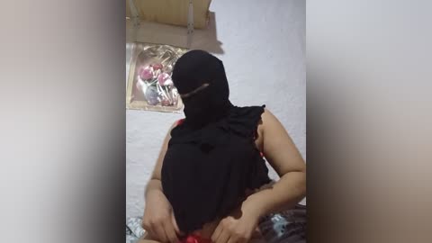 Media: Video of a woman in a black burqa, with a partially visible face, sitting in a room with white walls and a floral painting.