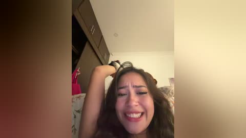 A video of an Asian woman with long black hair, wearing a red top, smiling and playfully pulling her hair. She's in a bedroom with a white ceiling and dark wooden furniture.