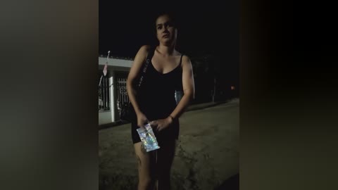 A dimly lit video of a woman standing on a street at night, wearing a black dress and holding a clear plastic bag. She has a serious expression.