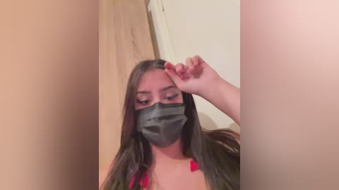 Video of a woman with long dark hair, wearing a gray face mask, touching her forehead, standing in a beige room with a wooden door.