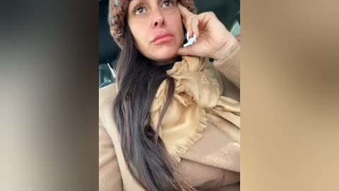 A video of a woman with long dark hair, wearing a brown knit beanie, beige scarf, and sweater, looking pensive and holding a phone to her ear.