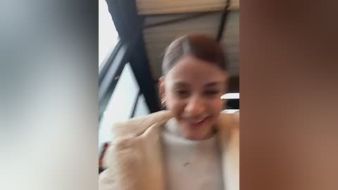 Video of a smiling young woman with light skin and brown hair in a bun, wearing a beige fur coat over a white sweater. Background shows a modern, industrial building with large windows.
