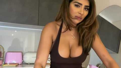 Video of a young woman with medium tan skin, long brown hair, wearing a low-cut black tank top, standing in a modern kitchen with grey cabinets and pink appliances.