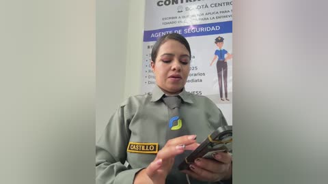 Video of a Latina woman with brown hair tied back, wearing a green uniform with a yellow badge reading \"INSPECCION LOCALES,\" inspecting a metal object in a sterile, white-walled room with a blue and white sign and a photo of a uniformed man.
