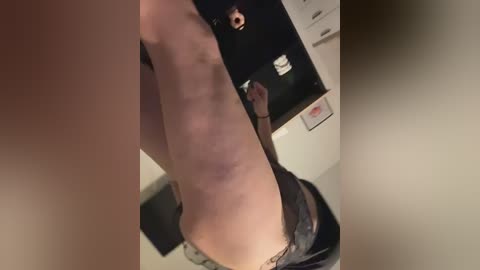 A video showing a woman with light skin, wearing black lace panties, standing in a dimly lit room. She is captured from a side angle, revealing her toned buttocks.
