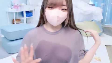 A video of an Asian woman with long brown hair, wearing a light gray t-shirt, a white face mask, and a white surgical mask, gesturing with her hands in a pastel-colored bedroom.