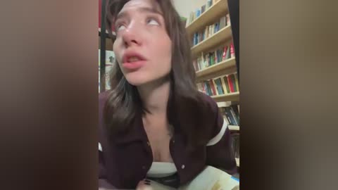 A video shows a Caucasian woman with shoulder-length brown hair, wearing a dark jacket, looking surprised. Behind her, a bookshelf is filled with colorful books.