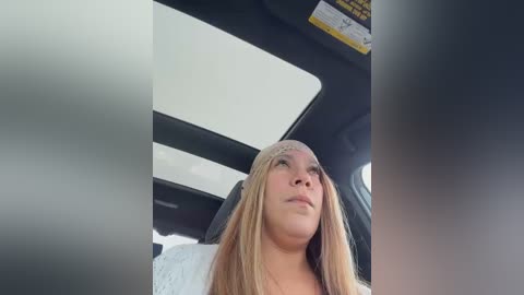 Media: Video of a blonde woman in a car, wearing a light sweater, looking up thoughtfully. The car's interior features a white dashboard and a yellow warning sticker.