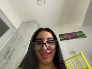 A video of a young woman with long black hair, glasses, and a wide smile, standing in a white room with a wooden bench and a colorful flower painting on the wall.