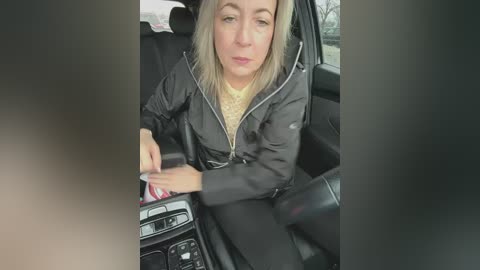 Media: Video of a blonde woman with fair skin, wearing a black jacket, driving a car; she holds a cell phone.