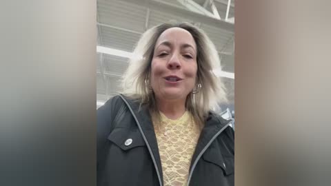 Video of a middle-aged woman with shoulder-length blonde hair, fair skin, and a warm smile, wearing a yellow lace top under a black jacket, indoors with a warehouse-like background featuring white ceiling panels and metal beams.