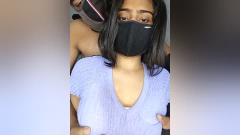 A video of a young woman with medium skin tone and straight black hair, wearing a black face mask and a light purple textured top, being held by a person's hands.