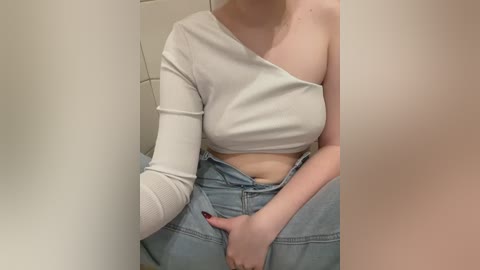 Video of a light-skinned woman with a slender physique, wearing a white, off-the-shoulder crop top and light blue high-waisted jeans. She sits with her legs slightly apart, one hand on her thigh, and the other partially covering her crotch. Background is a plain, off-white wall.