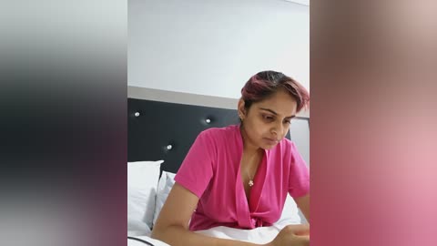 A video of a South Asian woman with medium brown skin, short hair, and a pink shirt, reading a book in a modern, minimalist bedroom with a black headboard.