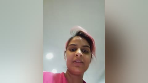 Video of a young woman with medium brown skin, wearing a pink top and headphones, looking directly at the camera, taken indoors with blurred background.