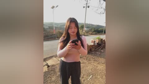 Video of an East Asian woman with long dark hair, wearing a pink long-sleeve top and black checkered pants, standing outdoors on a dirt path, holding a smartphone.