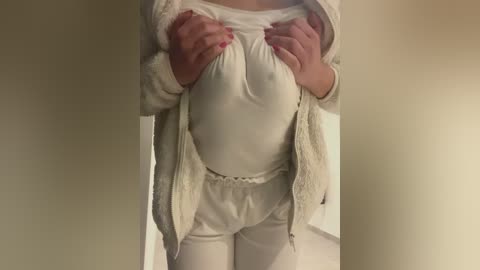 Video of a person with light skin, wearing a white, loose-fitting top and pants, lifting their top to reveal a large, round, pregnant belly. Hands with red-painted nails grip the shirt. Background is a plain, beige wall.