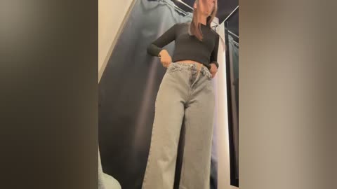 Video of a young woman in a dressing room, wearing a black long-sleeve top and high-waisted, light gray bell-bottom jeans. She stands confidently, hands on hips. Background features a black curtain and white garment on a hanger.