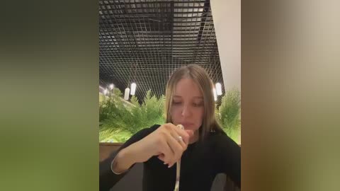 Video of a young Caucasian woman with light brown hair, wearing a black top, eating a donut in a modern, indoor setting with a black grid ceiling and green plants in the background.