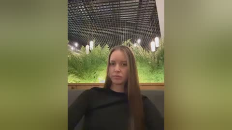 Video of a young woman with straight, long brown hair, wearing a black top, seated indoors with a modern, green-lit ceiling and lush green plants in the background.