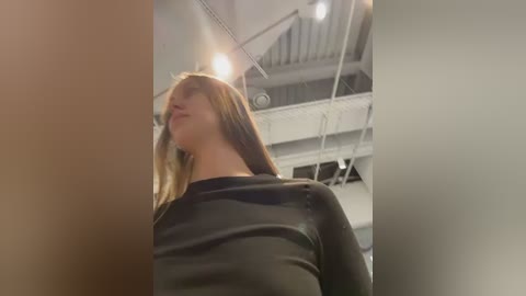 Video of a young woman with straight, long brown hair, wearing a black, long-sleeved top, standing indoors near a staircase with white railings and exposed metal beams.