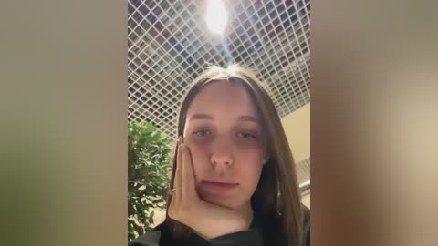 Video of a young Caucasian woman with long brown hair, resting her chin on her hand, in a room with a grid-patterned ceiling and a potted plant in the background.