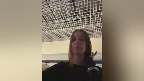 Video of a smiling woman with shoulder-length brown hair, wearing a dark top, standing under a grid-patterned ceiling in an indoor setting.