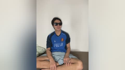 Media: Video of a young man with short dark hair, wearing sunglasses, a blue sports jersey, and gray shorts, sitting cross-legged on a bed against a white wall.