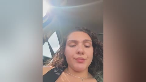 Video of a young woman with curly dark hair, wearing a black top, and a silver necklace, smiling inside a car with sunlight streaming through the windshield.