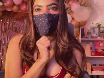 Media: Video of a young woman with long brown hair, wearing a navy blue mask with white polka dots, a red bra, and a playful expression. Background features pink balloons, a shelf with books, and a curtain.