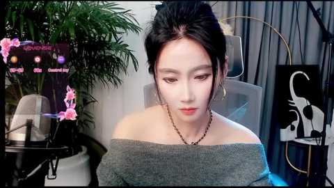 Video of an East Asian woman with fair skin, black hair styled in a bun, wearing a gray off-shoulder sweater, sitting in a modern chair with a green plant, pink flowers, and a black abstract painting in the background.