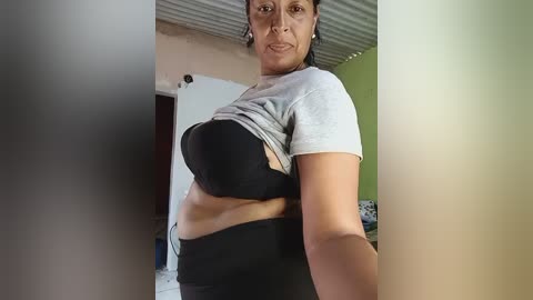 Video of a curvy Latina woman with medium skin tone, wearing a gray t-shirt and black bra, revealing her stomach. She stands in a simple room with a corrugated roof and light-colored walls.